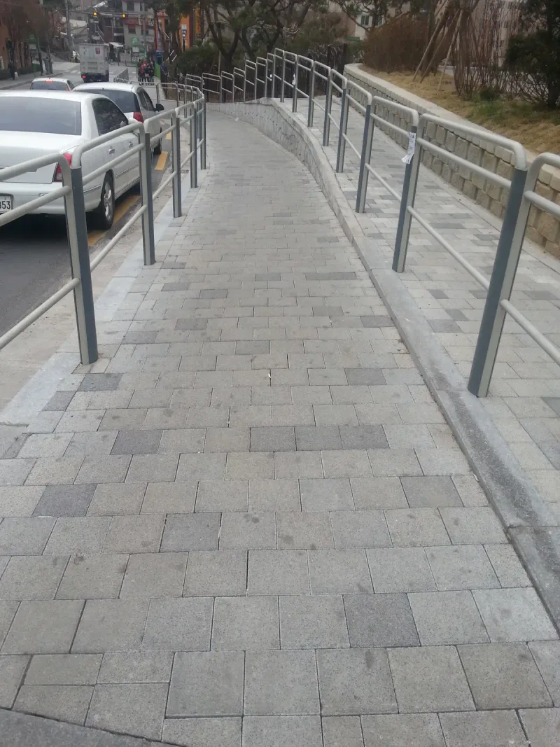 불광 롯데 현장 앞 경사 보도. 회색 직사각형 블록이 엇갈려 깔린 오르막 보도 양옆에 난간이 서 있고, 오른쪽 위 단에는 옹벽 블록 화단을 따라 또 하나의 보행로가 나란하다.