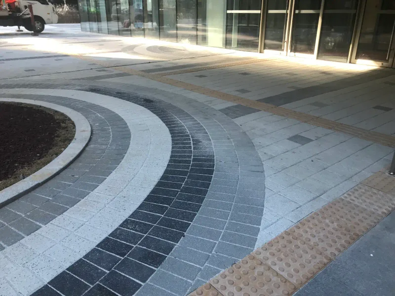 문정동 SK 건물 출입구 앞 바닥. 원형 화단을 따라 밝은 회색·짙은 회색·검은 블록 띠가 곡선을 그리고 옆에 노란 점자블록이 지난다.