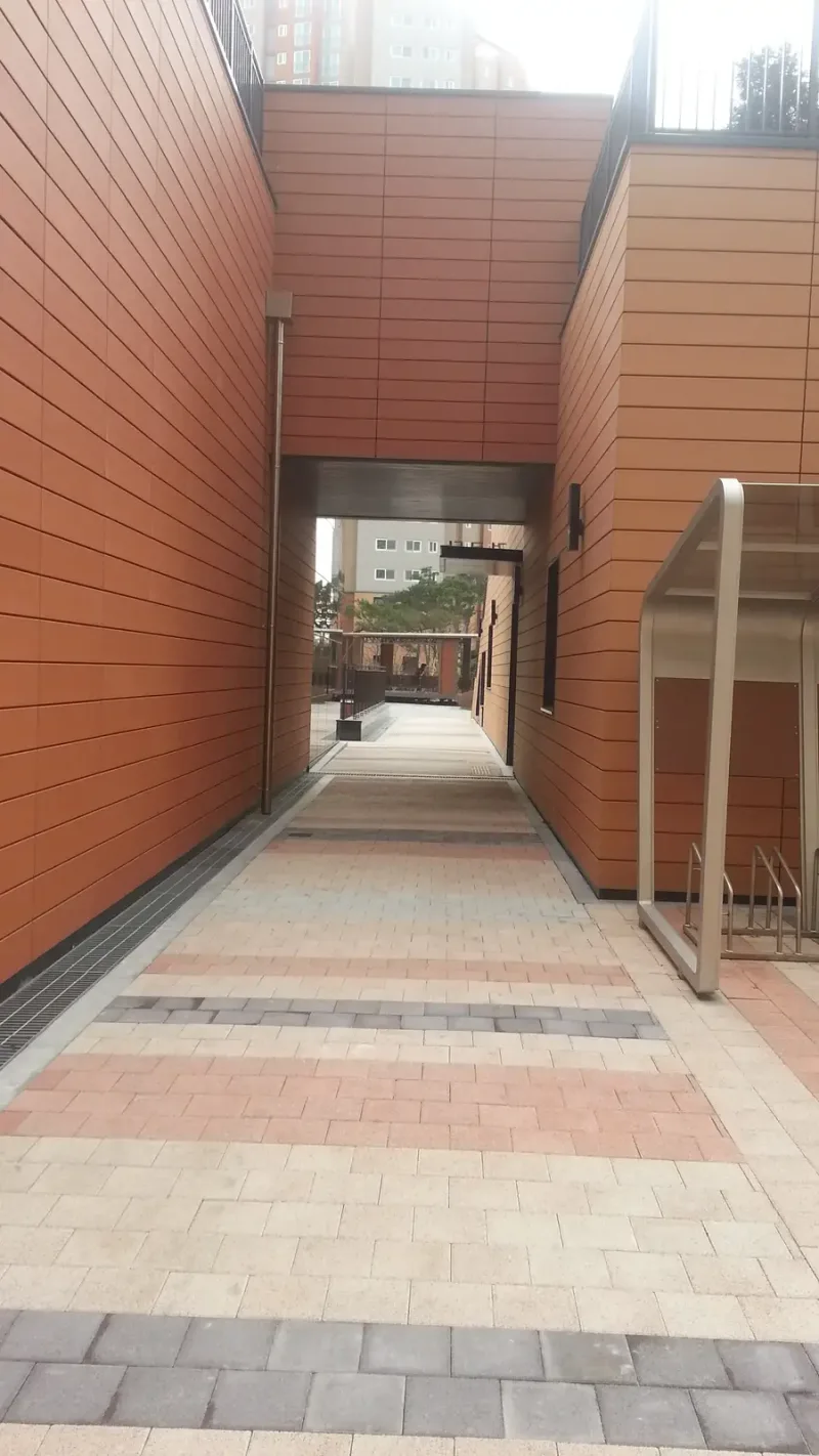 영통 래미안 마크원 동 사이 통로를 세로로 담은 모습. 베이지 블록 바닥에 분홍·짙은 회색 띠가 되풀이되며 건물 사이 필로티 너머로 이어진다.