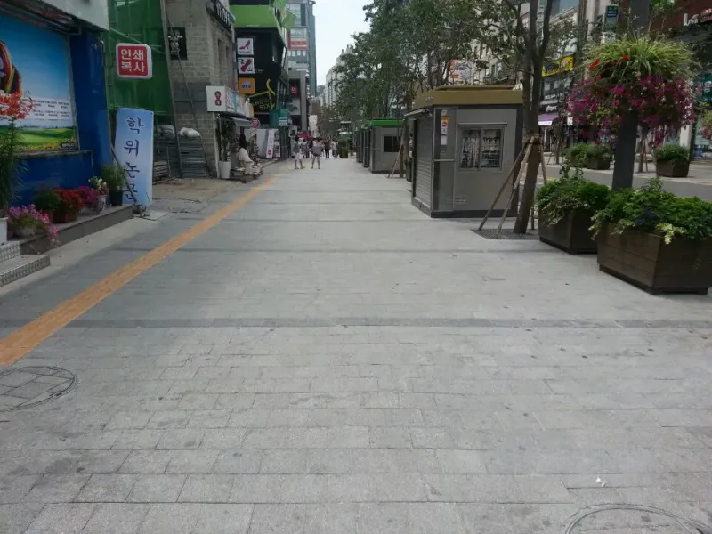 신촌 연세로 보행 공간을 멀리 내다본 모습. 회색 블록 거리 한쪽에 노란 점자블록, 다른 쪽에 꽃 화분과 가판대가 줄지어 있다.