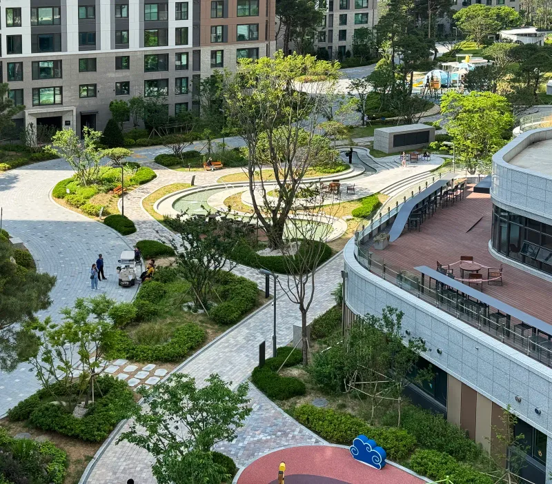 산성역 자이푸르지오 중앙 정원을 내려다본 모습. 회색 블록 산책로가 화단과 수경시설 사이로 굽이치고 옆에 커뮤니티 건물 데크가 있다.