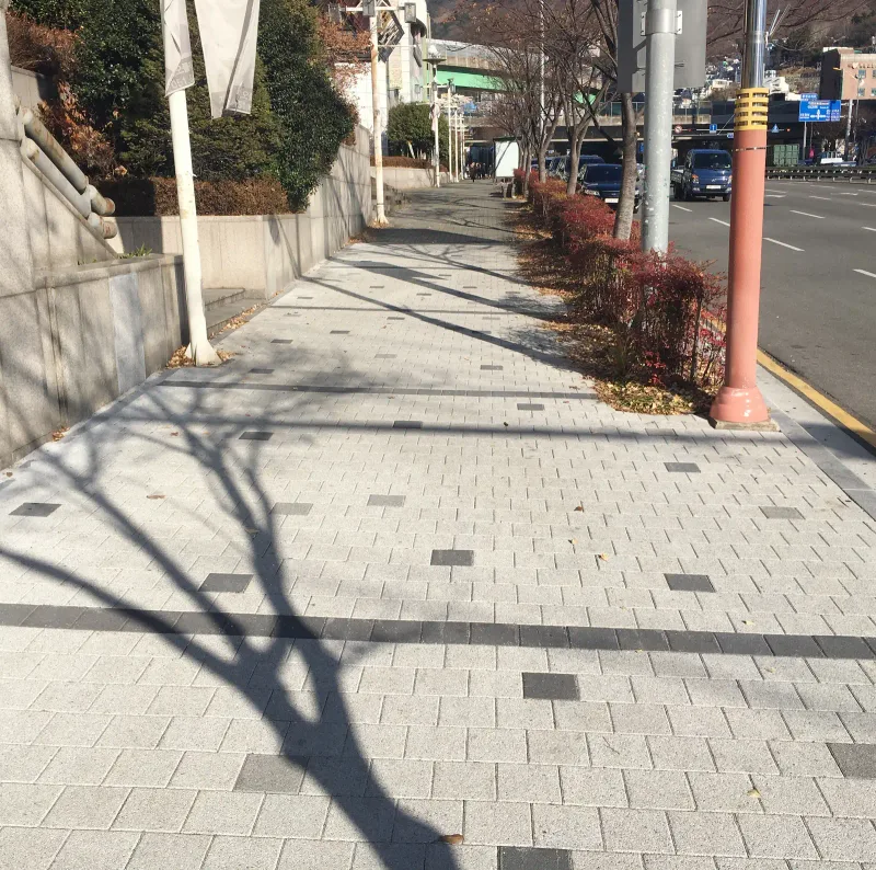 지오플레이스 일원 도로변 보도. 연갈색 블록 포장 위로 가로수 그늘이 드리워지고 붉은 기둥 보호대 너머로 차도가 나란히 놓여 있다.