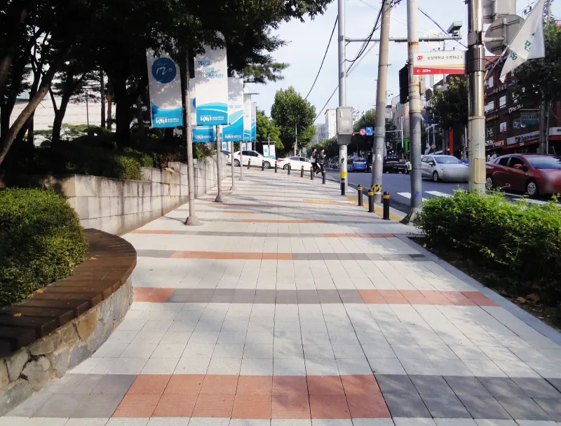 숭실대학교 중문 앞 보도. 밝은 회색 블록 바탕에 주황·짙은 회색 블록 띠가 가로로 반복되고 화단과 볼라드를 따라 길이 이어진다.
