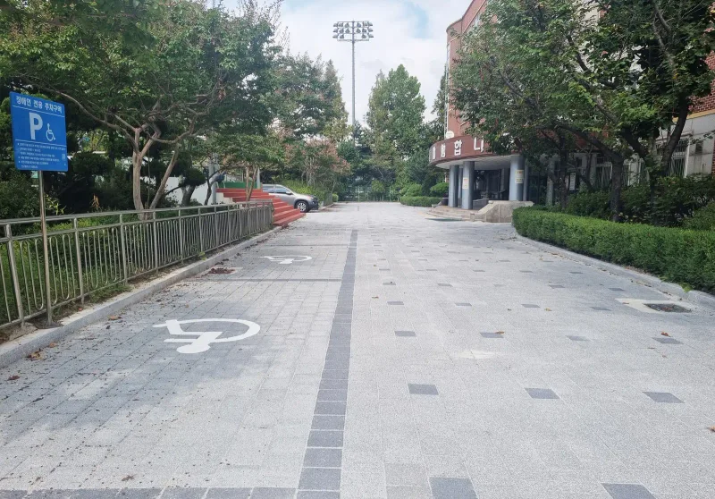 중원초등학교 진입로. 밝은 회색 블록 바닥에 짙은 회색 블록이 점과 띠로 놓여 있고 장애인 주차 표시와 교사 현관이 보인다.