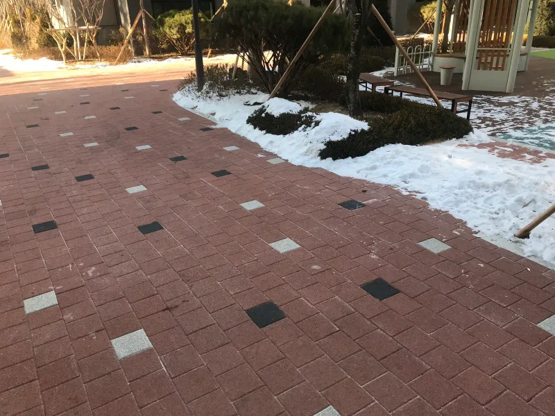 북한산 더샵 정자 앞 보행로. 적갈색 블록에 검은색·흰색 블록이 흩어진 길이 눈 쌓인 화단을 따라 휘어진다.
