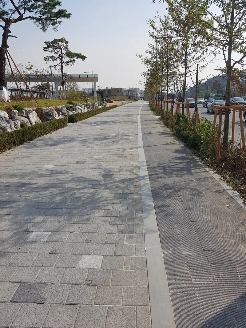 수서역 인근 가로수 보도. 연회색 블록 보도와 짙은 회색 블록 구간을 경계석이 나누며 곧게 뻗고 양옆에 어린 가로수와 자연석 화단이 있다.