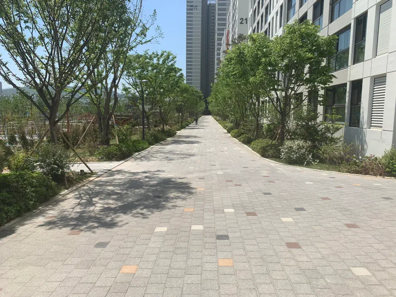 송파 헬리오시티 아파트 동 옆 넓은 보행로. 연회색 블록 바탕에 적갈색·주황·짙은 회색 블록이 점점이 섞여 있고 양옆에 나무가 있다.