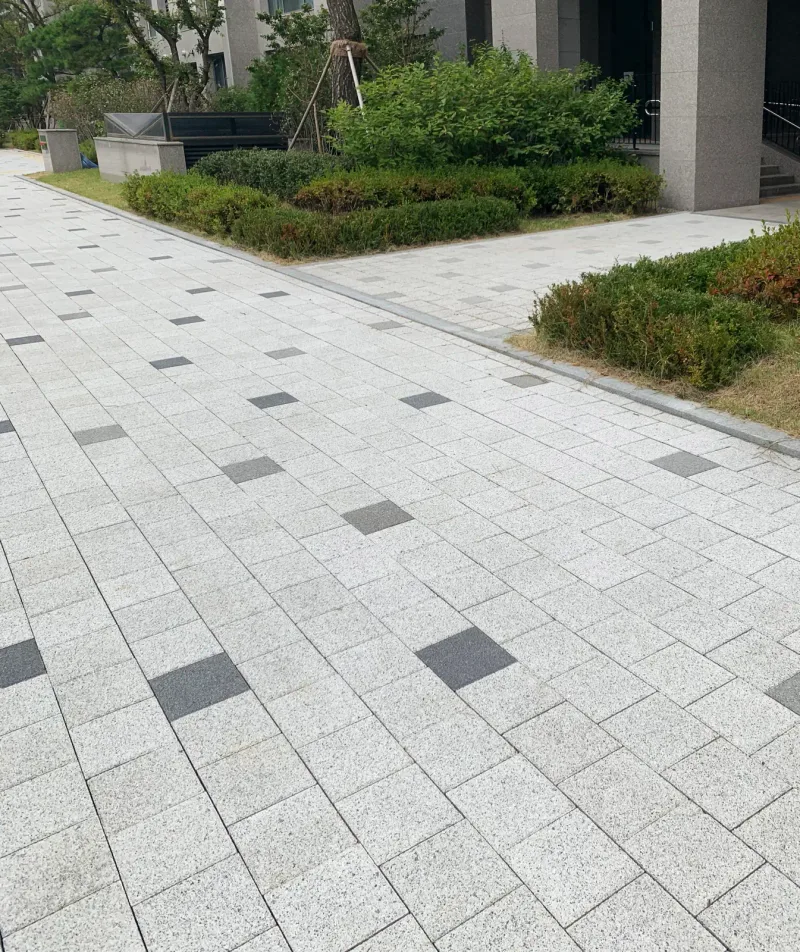 고덕 아르테온 동 입구 앞 보행로. 밝은 회색 블록 바탕에 짙은 회색 블록이 드문드문 박혀 있고 옆에 낮은 관목 화단이 있다.