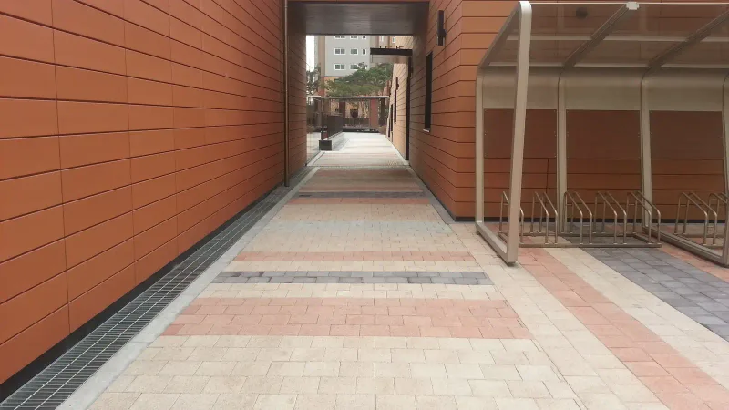 영통 래미안 마크원 동 사이 통로. 베이지 블록 바탕에 분홍·짙은 회색 블록이 가로 띠를 이루고, 벽 쪽 배수 덮개와 자전거 보관대가 나란하다.