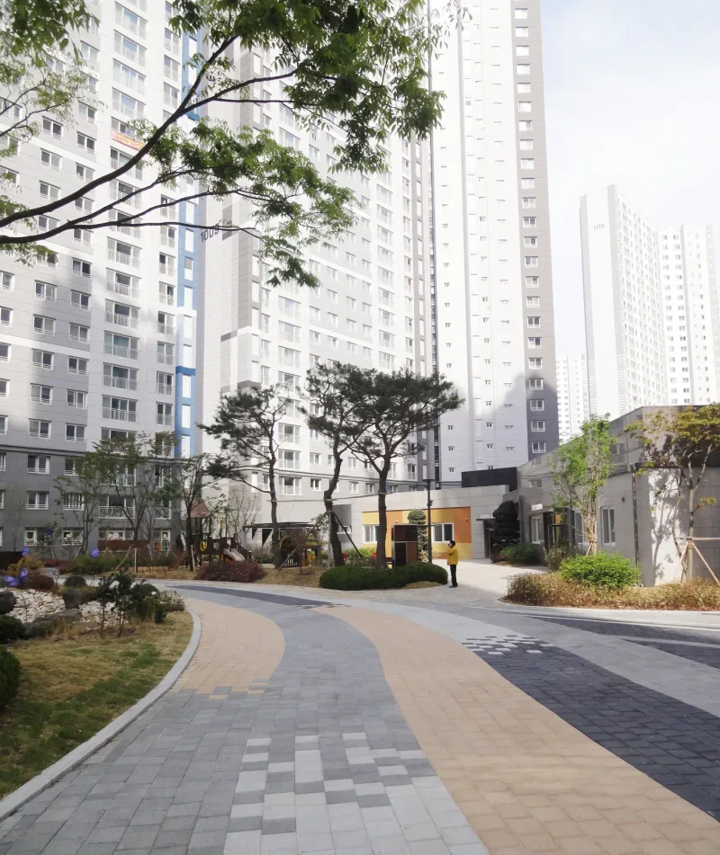 배곧 한신휴플러스 곡선 보행로. 회색 길에 베이지 띠와 진회색 띠가 휘어 흐르고 앞쪽은 흰색·회색 블록이 섞여 있으며 뒤로 소나무와 주민시설이 보인다.