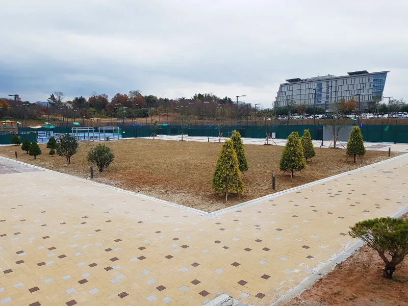 완주군 테니스장 진입 보행로 전경. 베이지 블록 바탕에 갈색·회색 블록이 점점이 박힌 넓은 길이 잔디 광장을 따라 꺾이고 뒤로 건물과 산이 보인다.