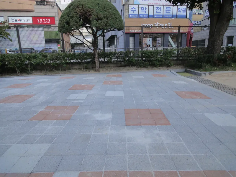 중계초등학교 운동장 끝 마당. 회색 블록 바닥에 적갈색·흰 블록이 사각 무늬로 섞여 있고 생울타리 너머로 상가 건물이 보인다.