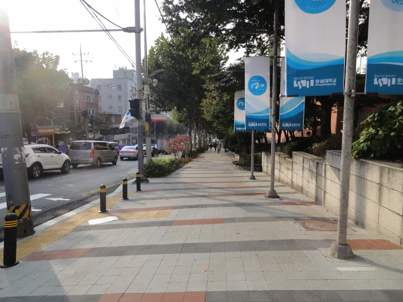 숭실대학교 중문 앞 교차로 쪽 보도. 적갈색·짙은 회색 가로 줄무늬가 들어간 연회색 블록 보도에 점자블록과 볼라드가 있고 가로등에 학교 배너가 걸려 있다.