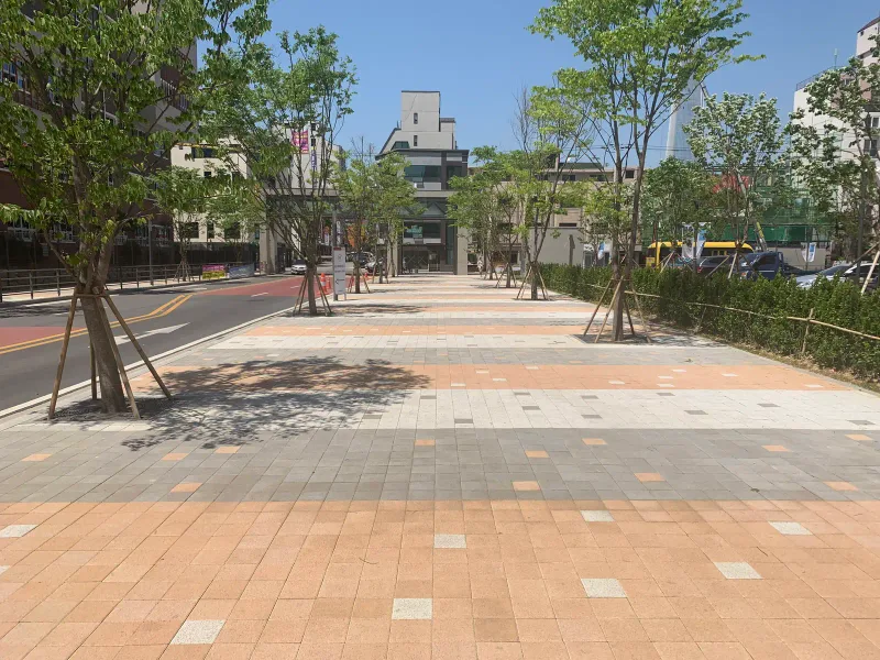 송파 헬리오시티 단지 앞 도로변 보도. 연적갈·회색 블록 띠가 가로수와 함께 넓은 보도를 따라 이어진다.