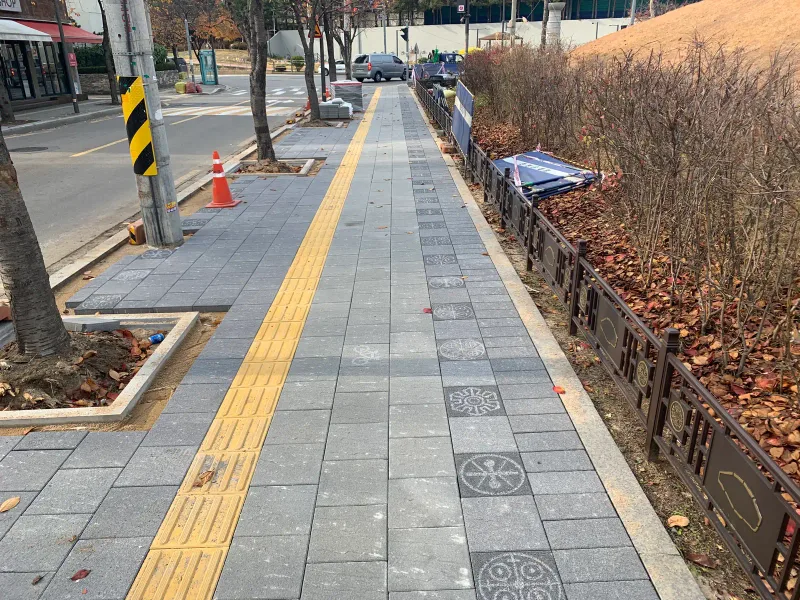 풍납토성 보도 원경(시공 마무리 중). 전통 문양 난간을 따라 회색 블록 보도와 노란 점자블록이 길게 뻗고 멀리 블록 더미와 고깔이 보인다.