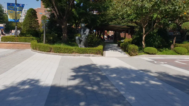 중앙대학교 조각상 정원 앞. 연회색·짙은 회색 블록 광장 위로 나무 그림자가 드리우고 뒤로 관목과 조각상이 보인다.