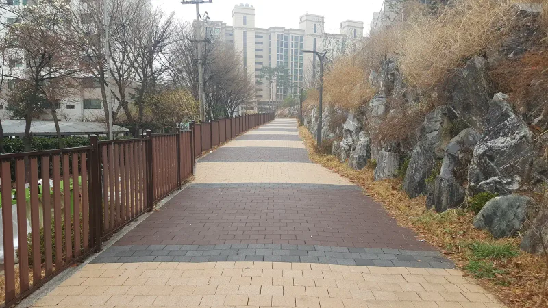 마포 현석 래미안 단지 옆 산책로. 목재 울타리와 바위 옹벽 사이로 베이지 블록 길이 뻗고 적갈색 블록이 넓은 띠로 가로질러 깔려 있다.