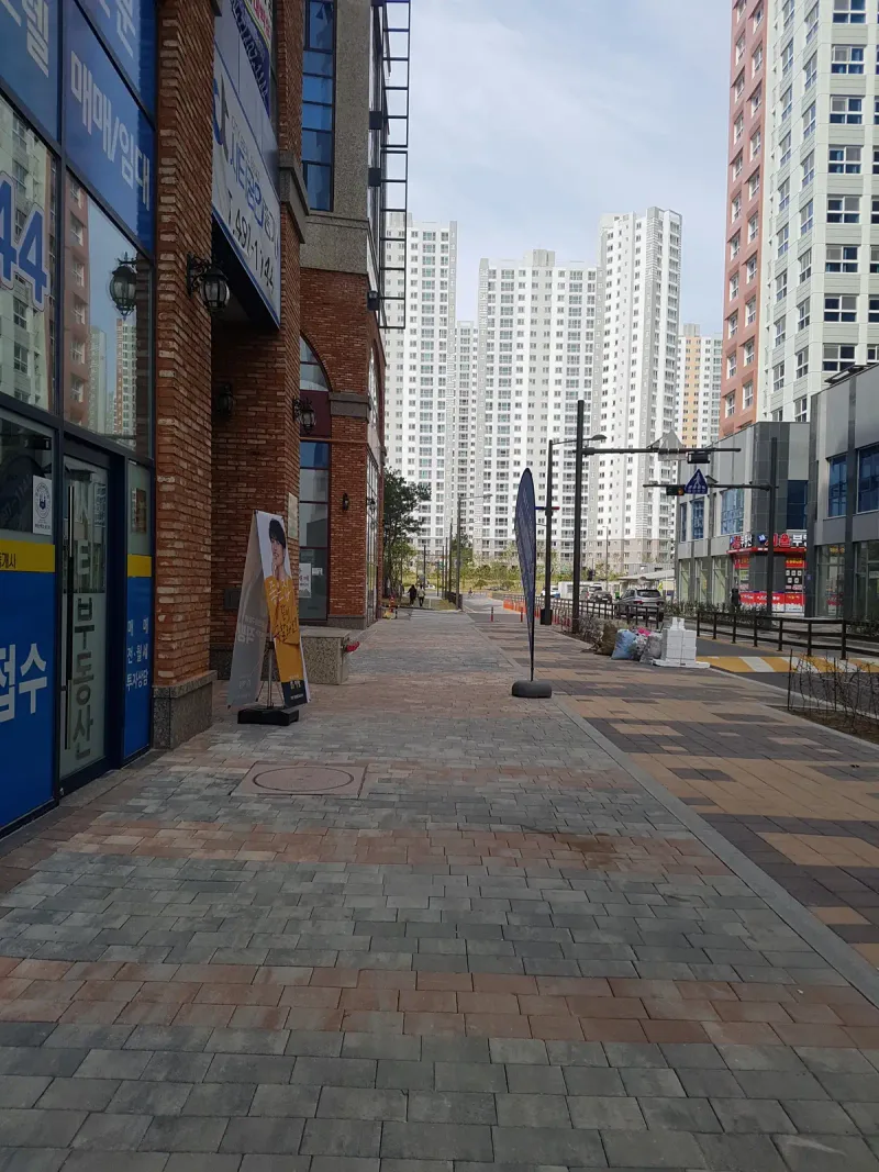 베니스스퀘어 상가 옆 곧은 보도. 회색 블렌딩 블록이 길게 깔려 있고 멀리 고층 아파트 단지가 보인다.