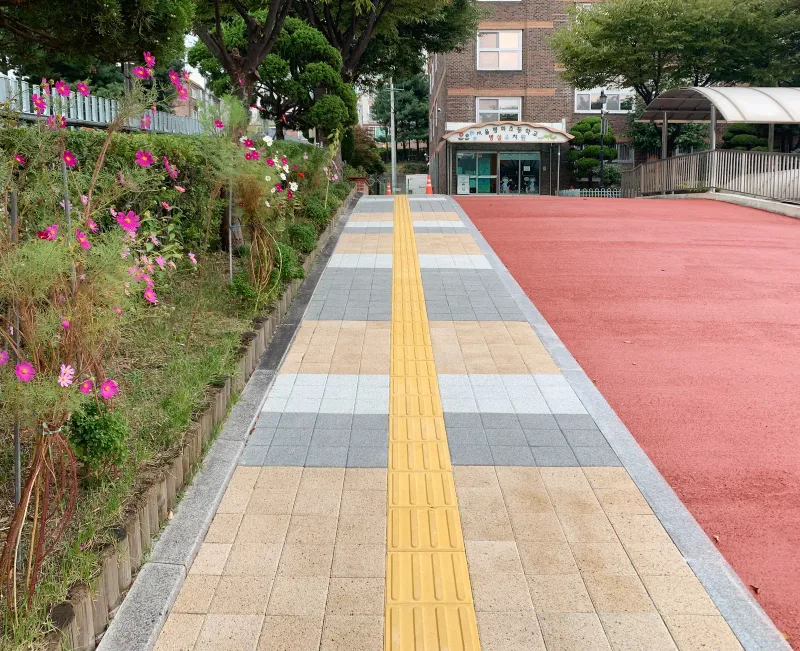 평화초등학교 병설유치원으로 이어지는 보행로. 베이지·회색·흰색 블록이 줄무늬로 깔리고 가운데 노란 점자블록이 놓였으며 오른쪽은 붉은 포장 진입로다.