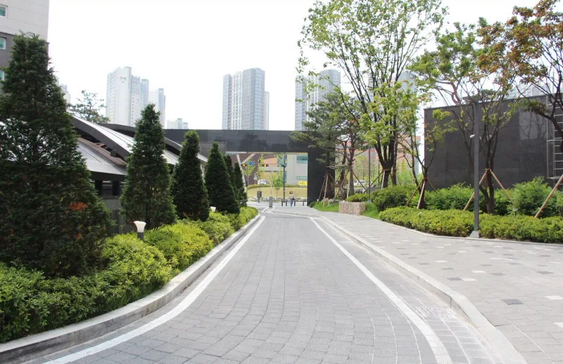 천안 불당 푸르지오 단지 내 곡선 통로. 회색 블록 길이 흰 경계석과 침엽수 화단을 따라 문주 쪽으로 휘어진다.