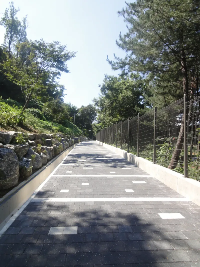 서울대공원 야구장 옆 숲길 세로 사진. 짙은 회색 블록 바닥에 밝은 블록 줄과 점이 반복되고 숲 사이로 길이 멀리 뻗는다.