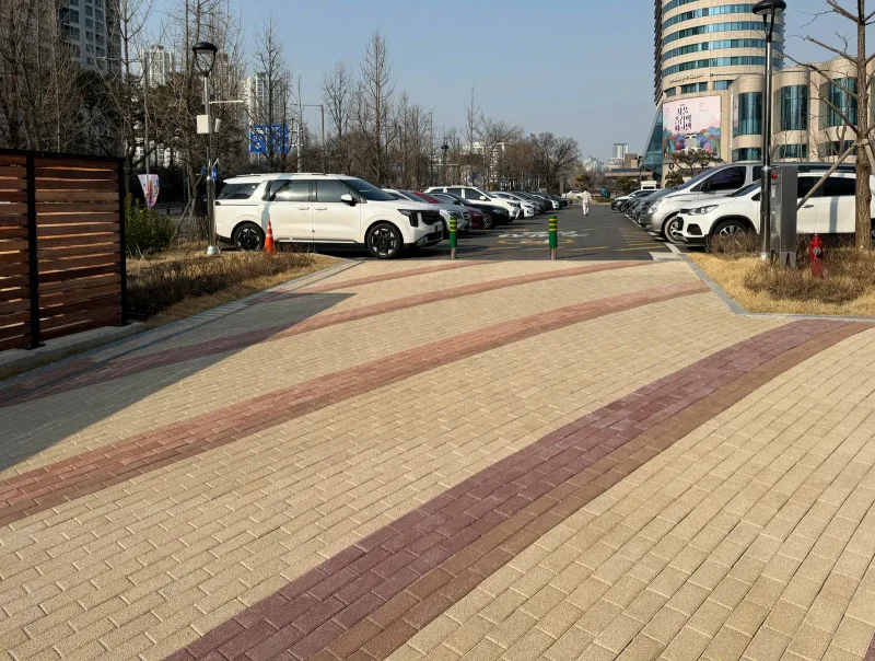 올림픽회관 앞 보행 광장. 베이지색 블록 바닥에 적갈색 띠가 완만한 곡선을 그리며 지나가고 뒤로 주차장과 건물이 보인다.