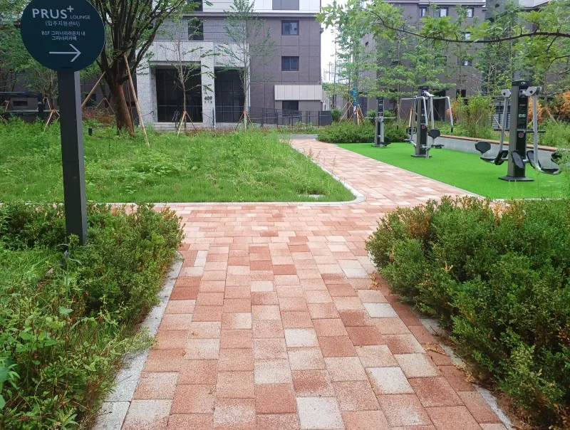 파주 운정 푸르지오 운동기구 쉼터 가는 길. 분홍·적갈색 블록이 깔린 보행로가 관목 사이로 이어지고 끝에 인조잔디와 운동기구가 있다.
