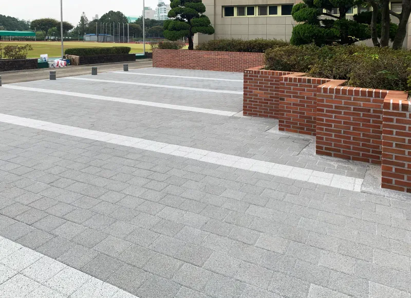 한국체육고등학교 운동장 쪽 마당. 회색 블록 바닥의 흰 줄무늬가 계단식 벽돌 화단 앞을 지나 멀리 운동장 방향으로 이어진다.