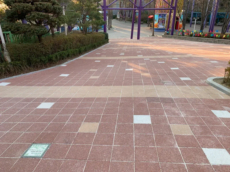 서울랜드 놀이시설 앞 보행로. 적갈색 정사각형 블록 길에 베이지 띠와 흰 블록이 들어가 있고 뒤로 놀이기구와 조경수가 보인다.