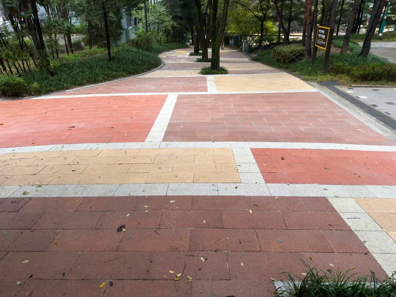상암동 매봉산로 산책로. 적색과 베이지 판형 블록 구획이 밝은 회색 띠를 사이에 두고 번갈아 이어지며 숲길로 뻗어 간다.