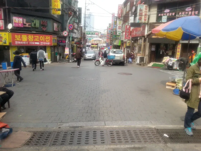 부천 소사 테마거리 시장 골목. 회색 직사각형 블록이 차도와 보행 공간 구분 없이 넓게 깔린 거리 양옆으로 상점 간판이 늘어서 있고, 멀리 테마거리 입구 아치가 보인다.