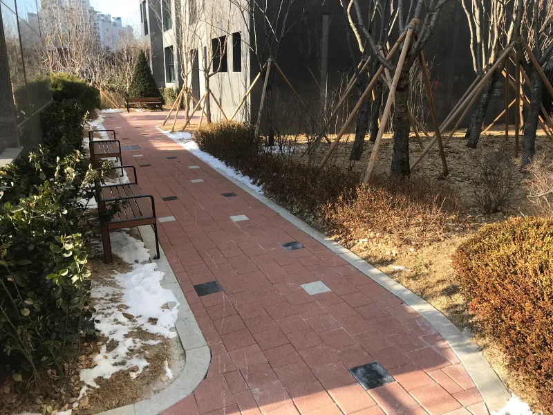 북한산 더샵 정원 산책로. 적갈색 블록 길이 경계석을 따라 굽어지고 한쪽에 나무 벤치가 이어진다.
