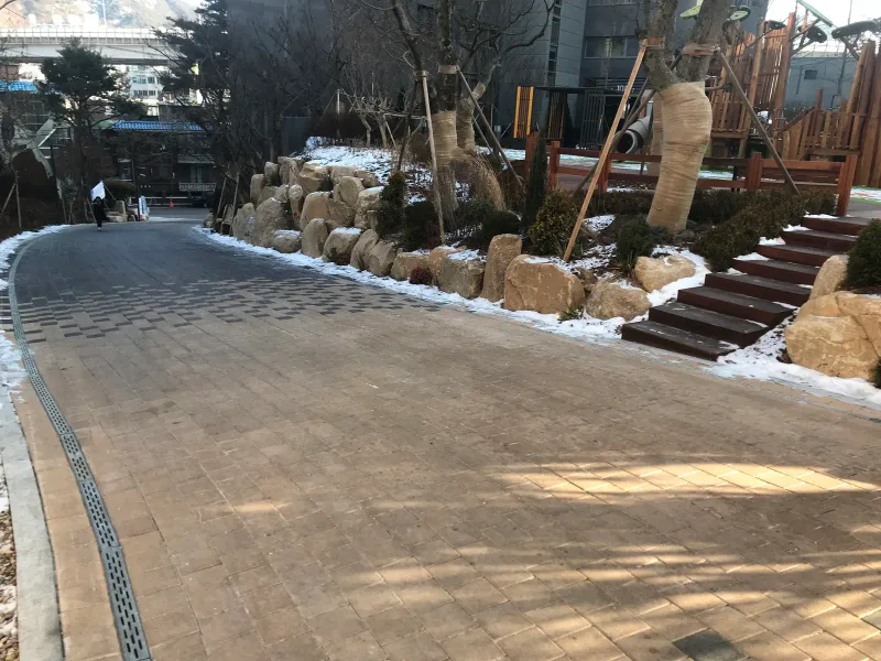 북한산 더샵 계단 옆 진입로. 베이지 톤으로 밝아지는 블록 길 옆으로 자연석 조경과 나무 계단이 놓여 있다.