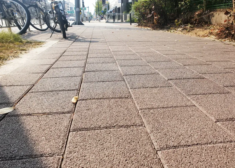강동 선사이야기길 자전거 거치대 옆 보도 근접. 갈색 블록의 거친 표면과 가지런한 줄눈이 햇빛 아래 뚜렷하게 보인다.