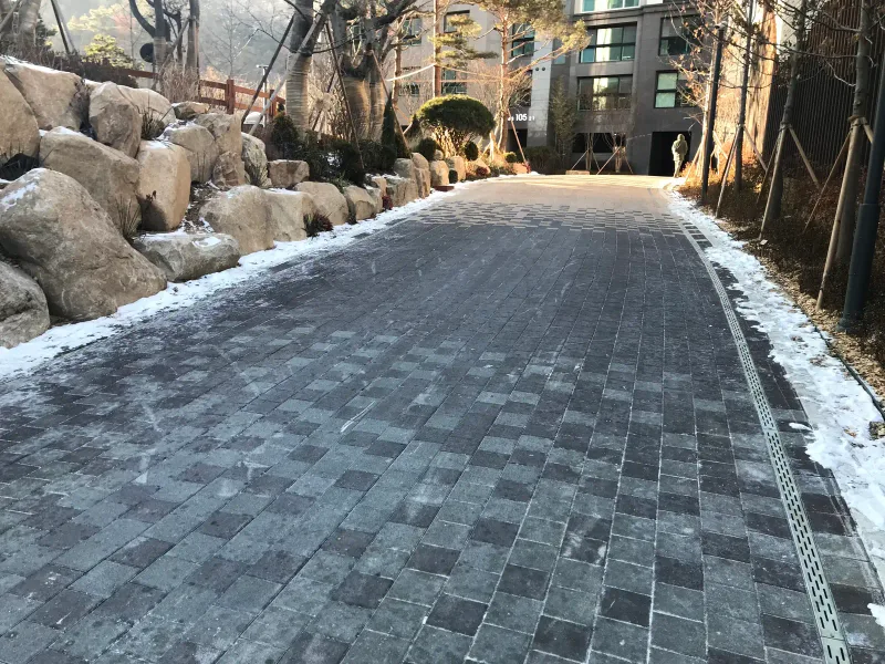 북한산 더샵 자연석 옹벽 사이 길. 짙은 회색과 연회색 블록이 섞인 넓은 포장면이 배수로를 따라 곧게 올라간다.