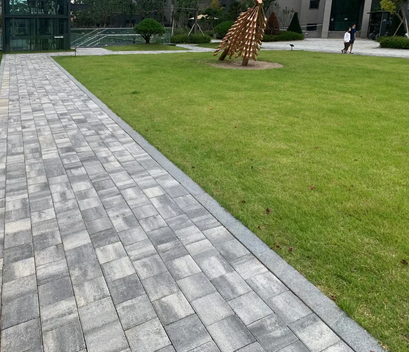 고덕 아르테온 넓은 잔디마당 가장자리 산책로. 여러 회색이 섞인 블록 길 옆으로 잔디와 조형물, 동 입구가 보인다.
