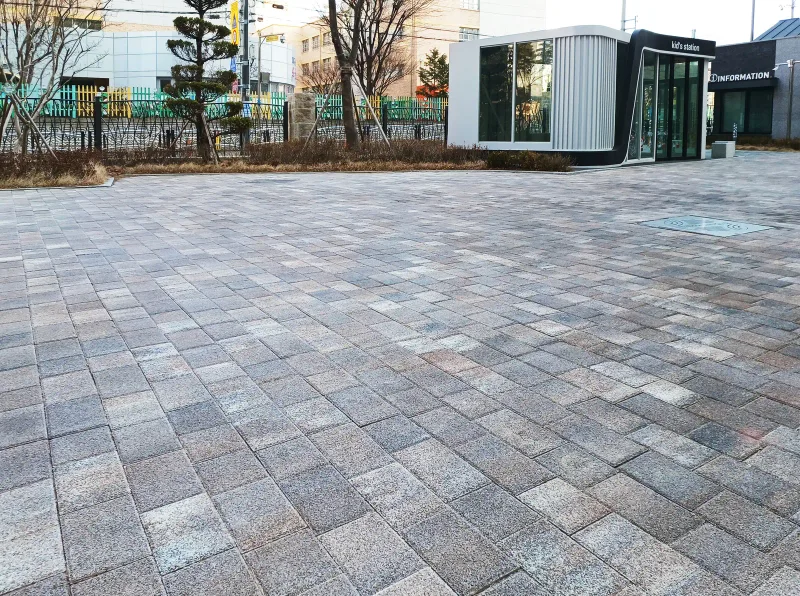 대구 달서 삼정그린코아 안내동 앞 마당. 회색과 갈색 톤이 섞인 블록이 넓게 엇갈려 깔려 있고 뒤로 울타리와 수목이 있다.