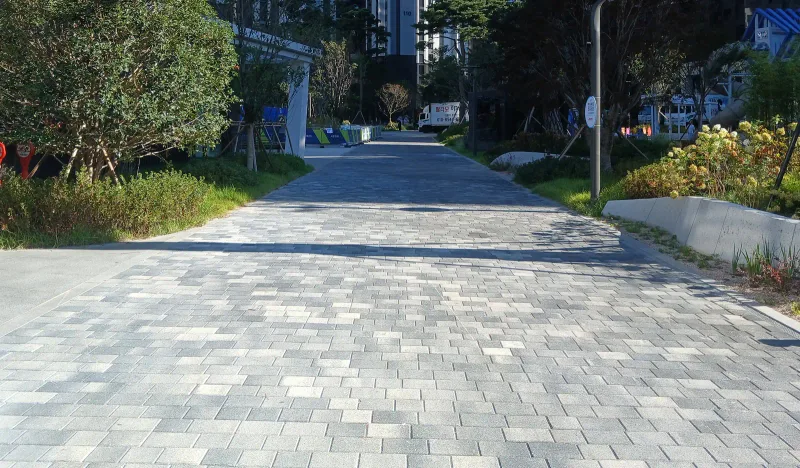 반포 래미안 트리니원 나무 사이 보행로. 밝은 회색 계열 블록이 넓게 깔려 있고 오른쪽 낮은 옹벽 위로 꽃과 관목이 심겨 있다.