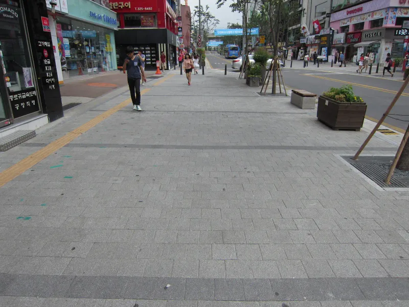 신촌 연세로 보행 공간. 밝은 회색 직사각형 블록이 넓게 깔린 거리를 짙은 회색 블록 가로 띠가 일정 간격으로 가르고, 상가 쪽으로 노란 점자블록, 차도 쪽으로 화분과 벤치가 놓여 있다.