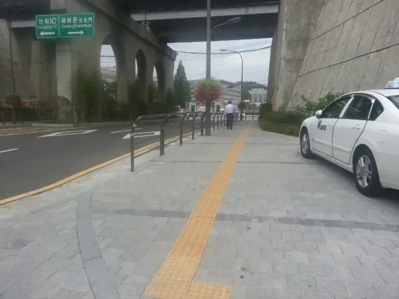 신촌 연세로 끝 철길 교각 아래 보도. 회색 블록 보도 가운데로 노란 점자블록이 곧게 뻗고, 곡선 경계석과 보행 난간이 차도와 나눈다.