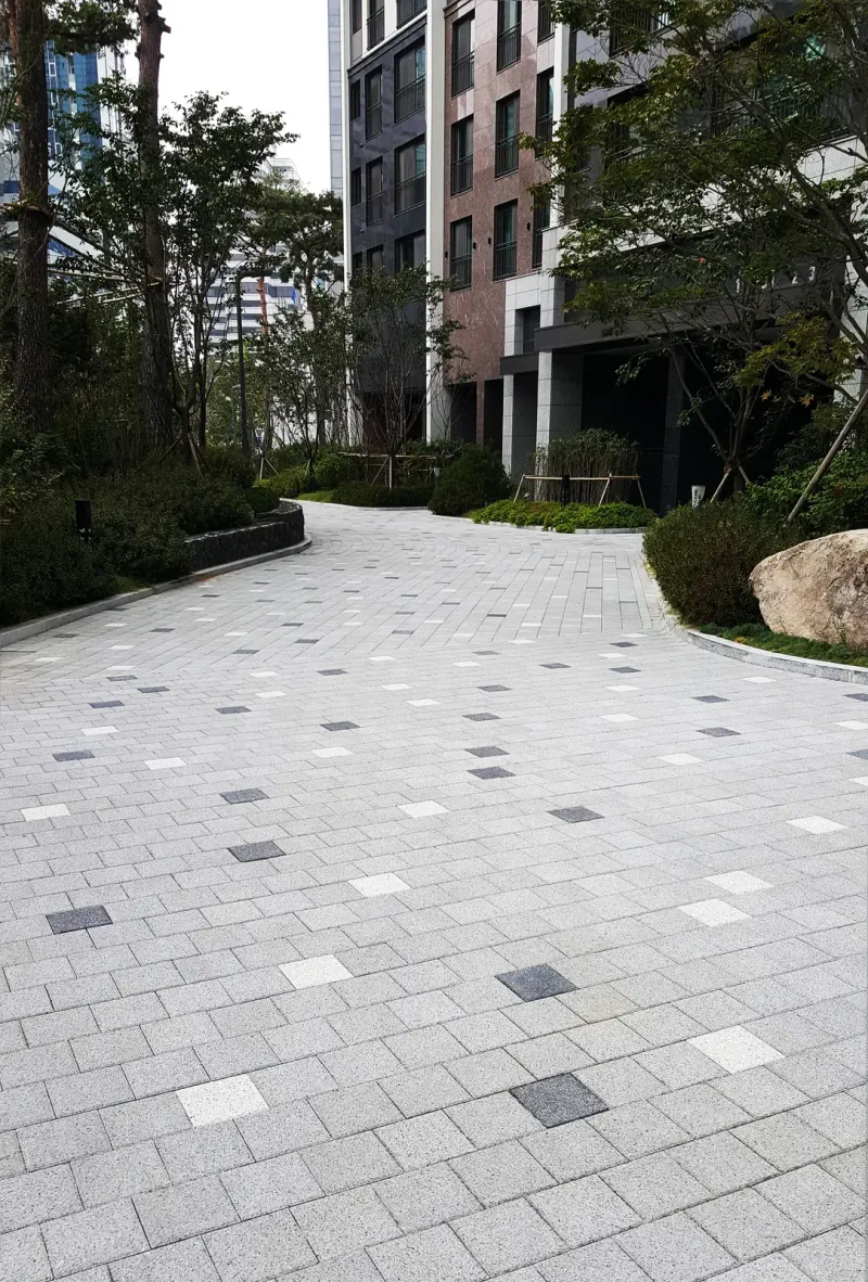 역삼 자이 단지 내 곡선 보행로. 연회색 직사각형 블록 바닥에 짙은 회색·흰색 블록이 흩어져 박혀 있고 길이 화단과 조경석을 끼고 아파트 동 앞으로 굽어 간다.