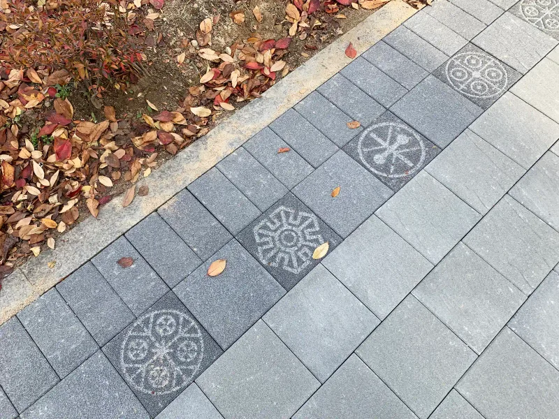 풍납토성 보도 문양 근접. 낙엽 진 화단 옆 회색 블록 줄에 바퀴·꽃 모양 등 서로 다른 옛 문양 블록이 번갈아 박혀 있다.