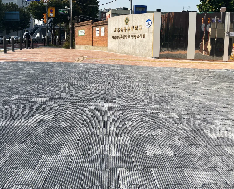마포 중동초등학교 교명 담장 앞 바닥. 흰 줄결이 들어간 회색 블록이 물결 모양으로 맞물려 깔려 있고 뒤로 분홍 블록 보도와 볼라드가 보인다.