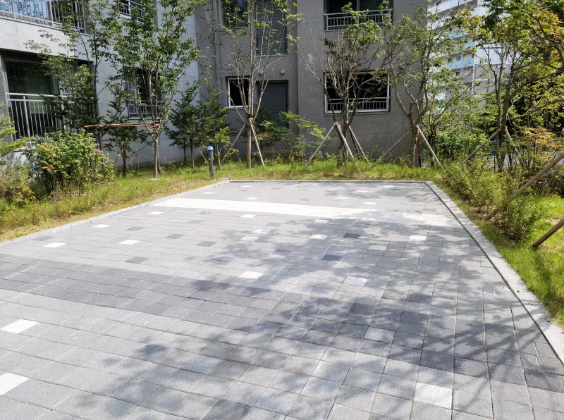 더샵 동천 이스트포레 동 앞 쉼터 포장면. 회색 블록 바탕에 흰 블록 띠와 짙은 회색 블록 줄이 들어가 있고 둘레에 잔디와 나무가 있다.