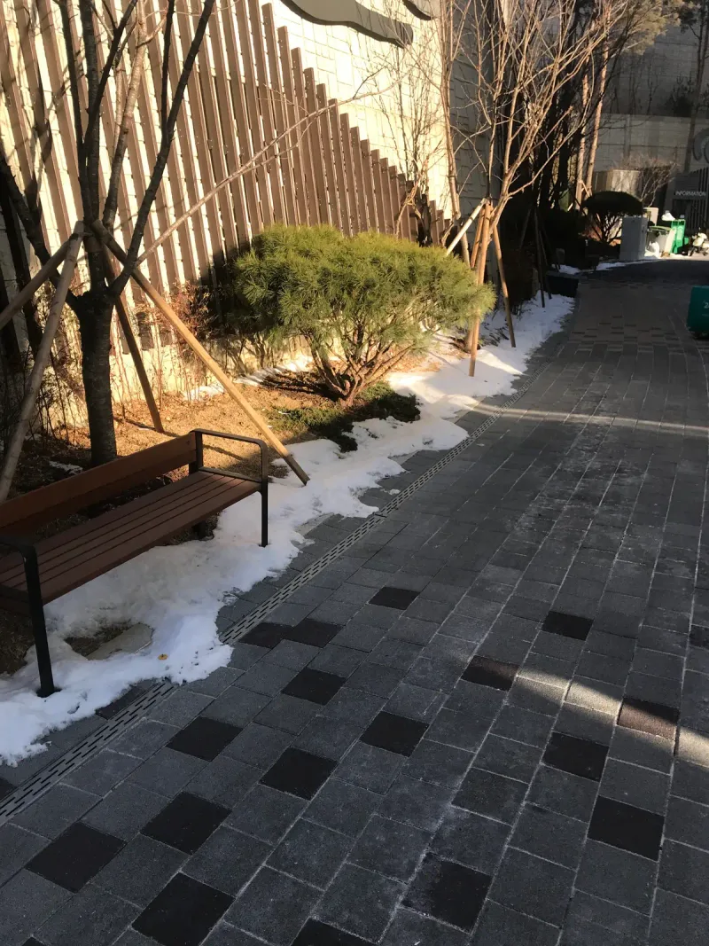 북한산 더샵 건물 옆 통로. 짙은 회색 바탕에 연회색 블록이 섞인 좁은 길과 나무 벤치를 세로로 담았다.