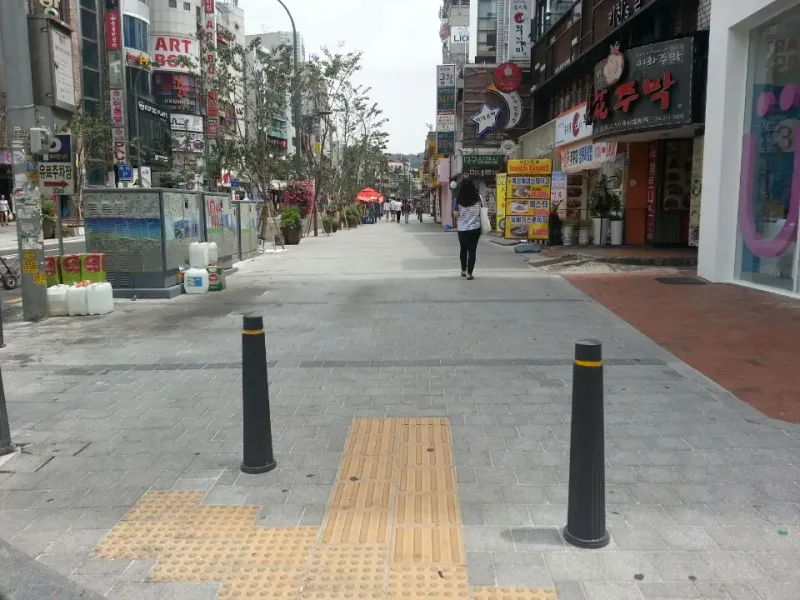 신촌 연세로 보도 진입부. 회색 블록 보도 입구에 검은 볼라드와 노란 점자블록이 놓이고, 상가를 따라 보도가 길게 이어진다.
